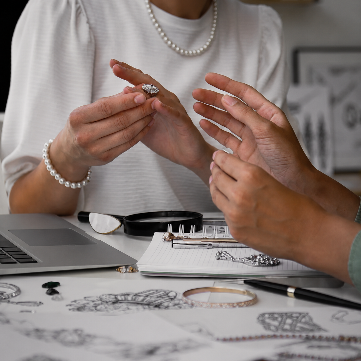 Two people examining jewelry at a desk with a laptop and magnifying glass.