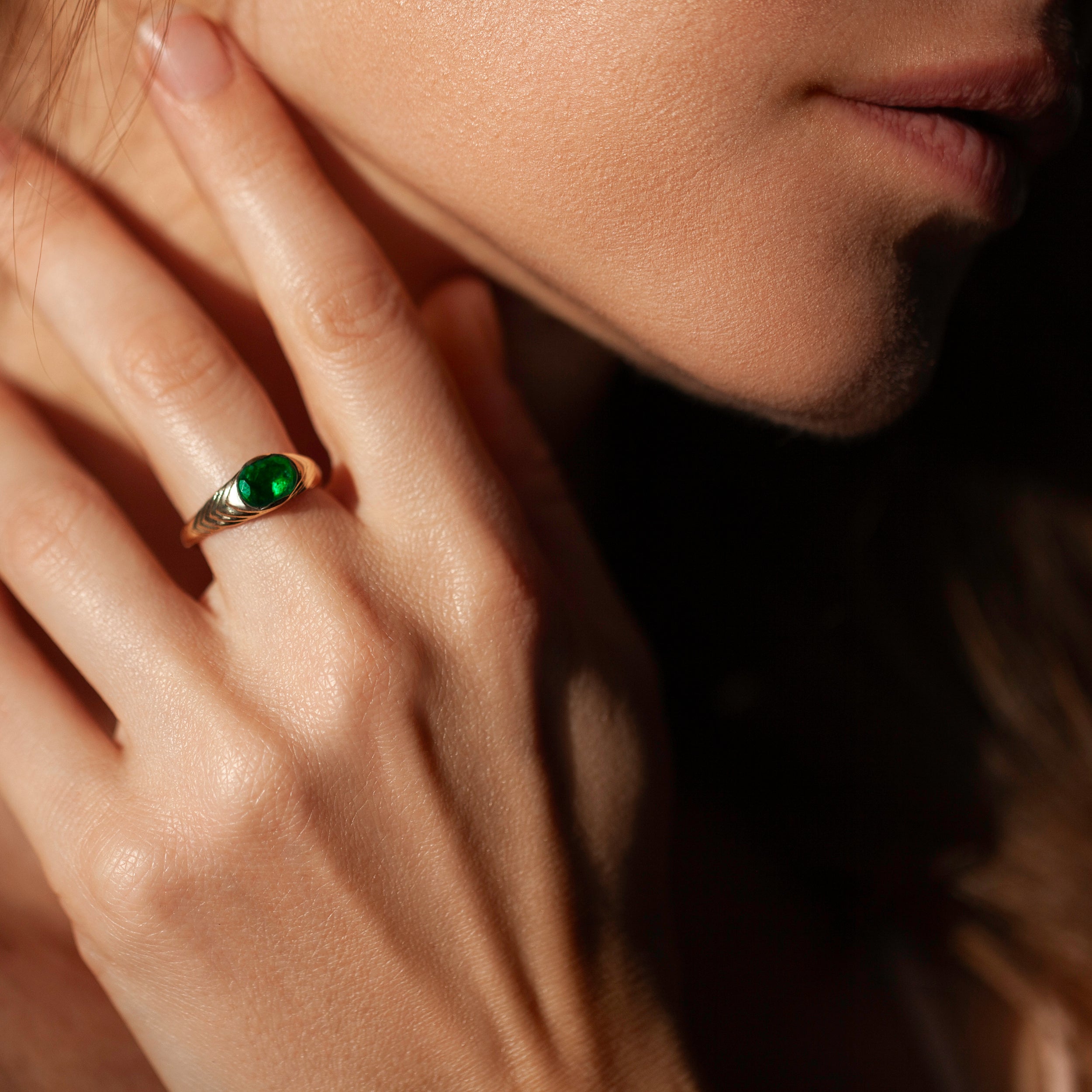 A close-up lifestyle shot of a model's hand wearing a gold ring with a prominent oval green gemstone, set against a dark, shadowed background.
