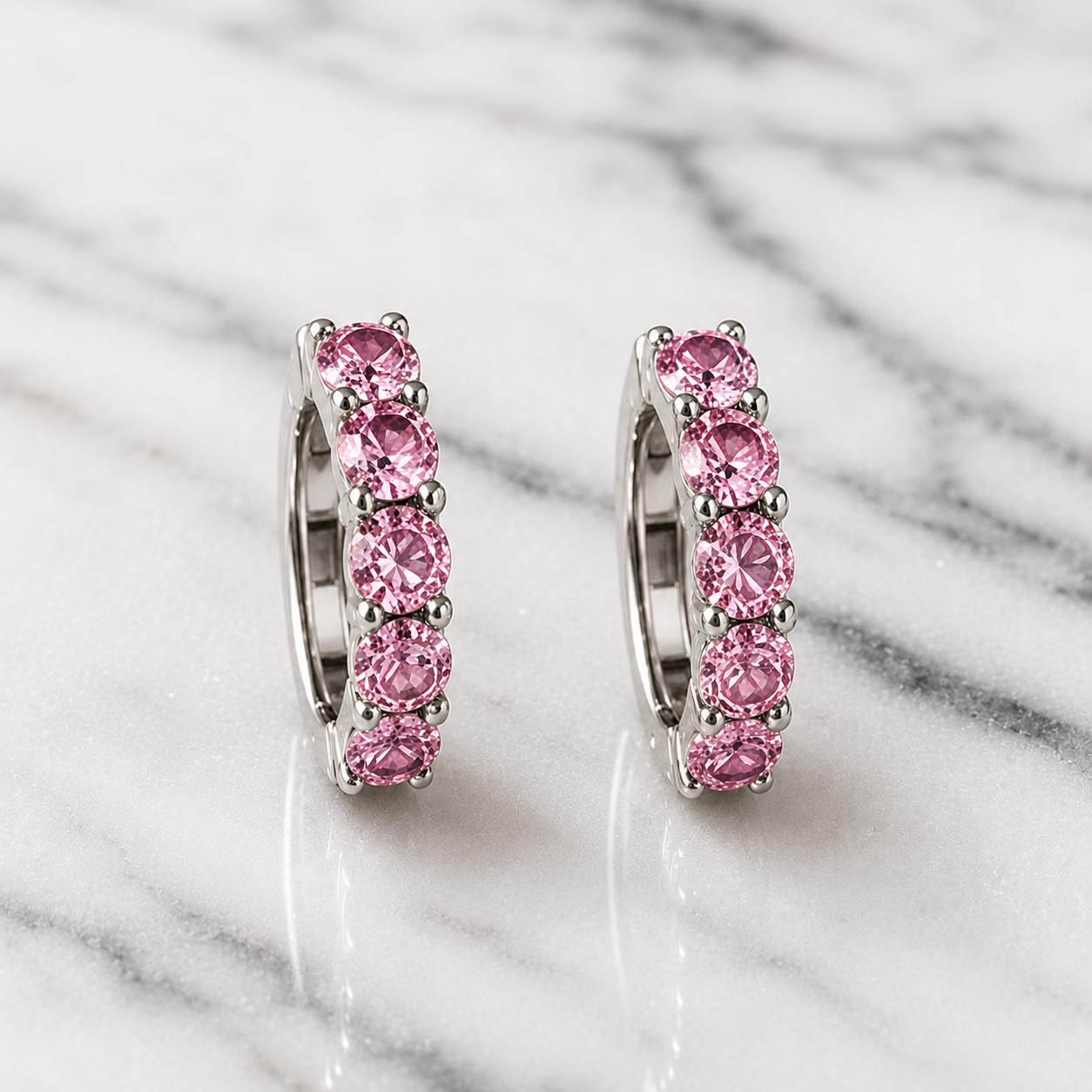 A pair of silver huggie hoop earrings set with five round pink gemstones each, displayed on a white and gray marble surface.
