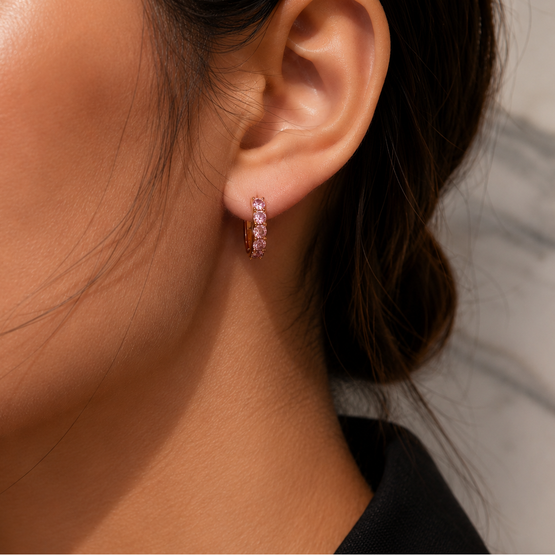 A close-up of a model's ear wearing a small pink hoop earring with clear stones, set against a soft gray marble background.