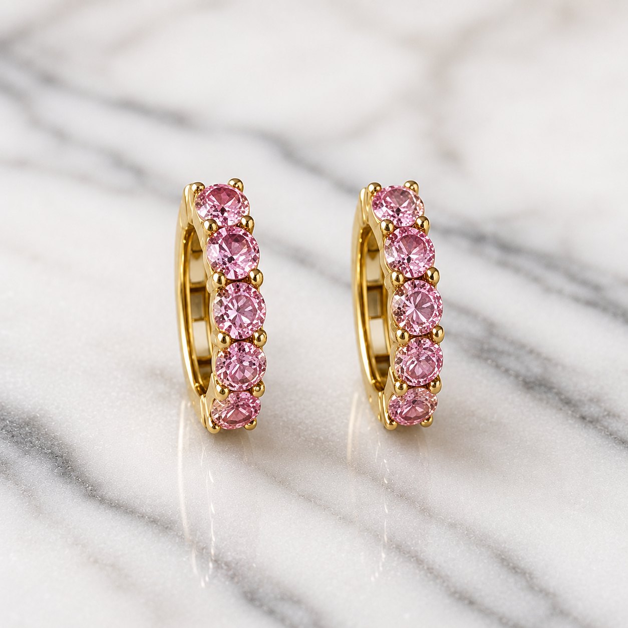 A pair of gold huggie hoop earrings set with round pink gemstones, displayed on a white and gray marble surface.