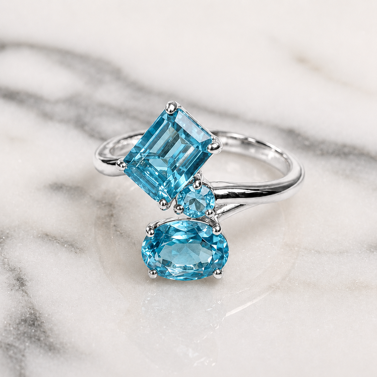 A silver ring featuring a large emerald-cut light blue gemstone, a smaller round blue stone, and an oval light blue stone, displayed on a white marble surface.