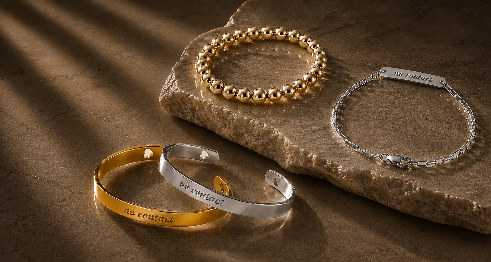 Three engraved bracelets displayed on a stone slab: a gold beaded bracelet, a silver chain bracelet with a rectangular tag, and a gold-and-silver cuff bracelet. The bracelets are photographed in warm directional light on a brown surface.