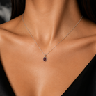 A close-up of a model's neck and chest wearing a delicate silver chain necklace with a small oval pendant featuring a dark purple stone.