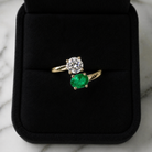 Gold ring with a diamond and emerald on a black cushion against a marble background