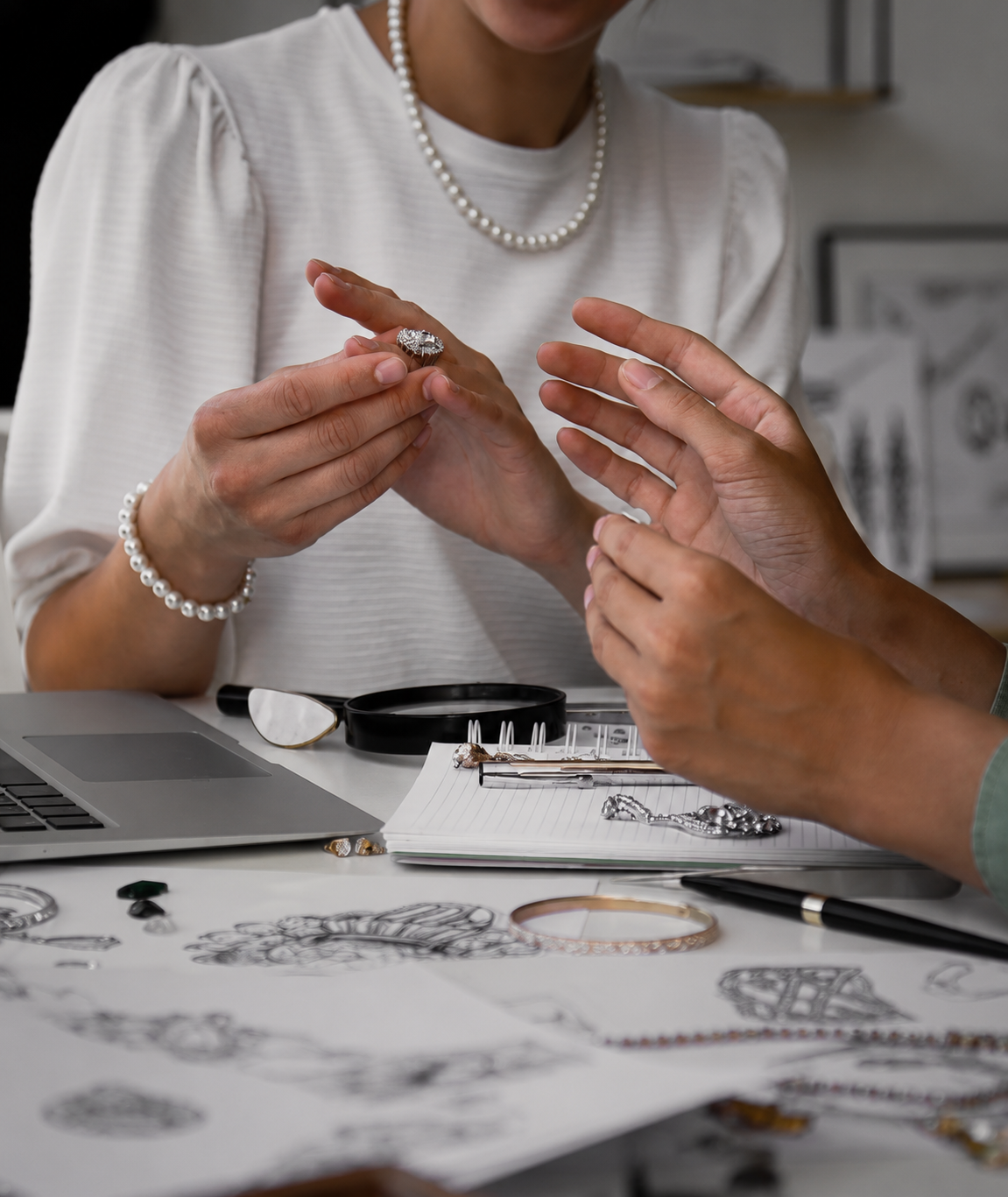 Two people examining jewelry at a desk with a laptop and magnifying glass.