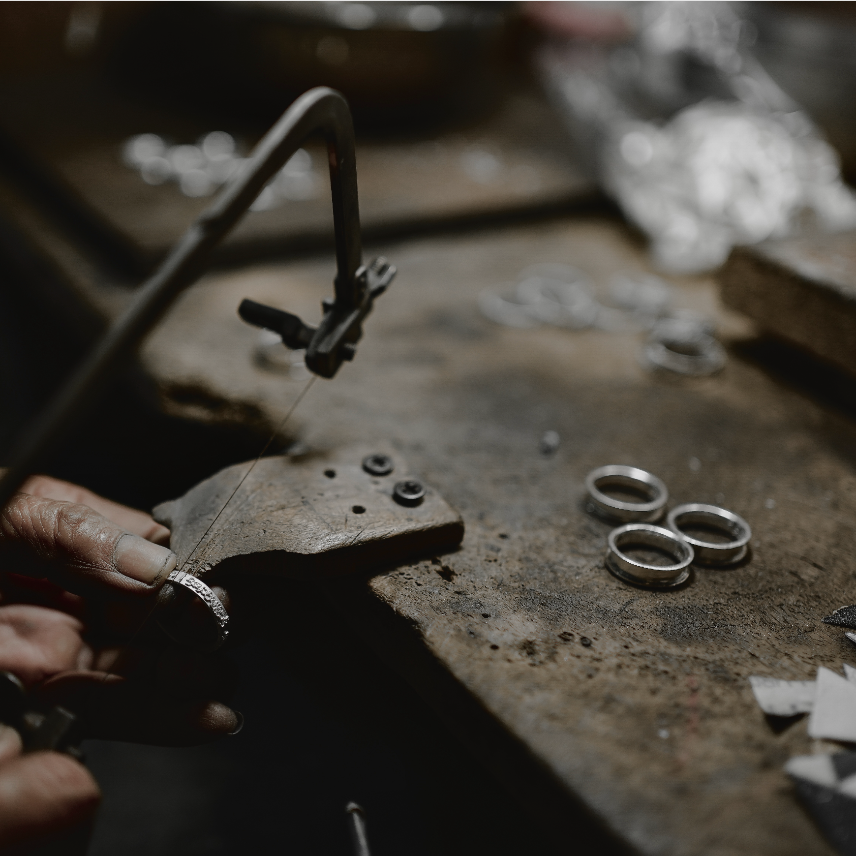A close-up of a jeweler's hands using a ring press and bowie saw to work on a ring at a rustic, dark workbench with other metal rings and crafting tools nearby.