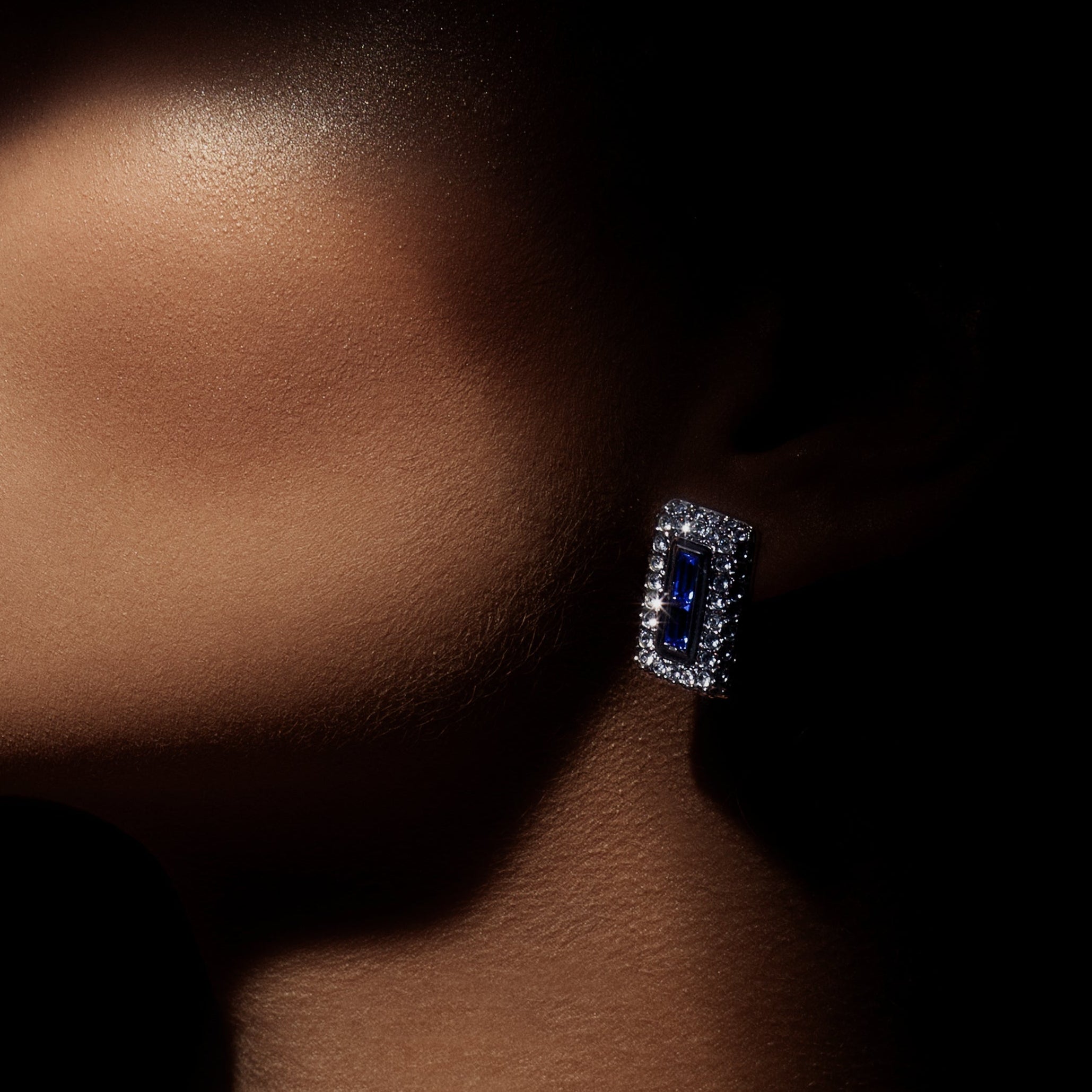 A close-up portrait of a woman wearing a silver-toned necklace and matching rectangular stud earring, both set with blue stones and clear pavé accents, against a dark background.