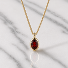 A gold chain necklace with an oval red gemstone pendant, displayed on a white marble surface.