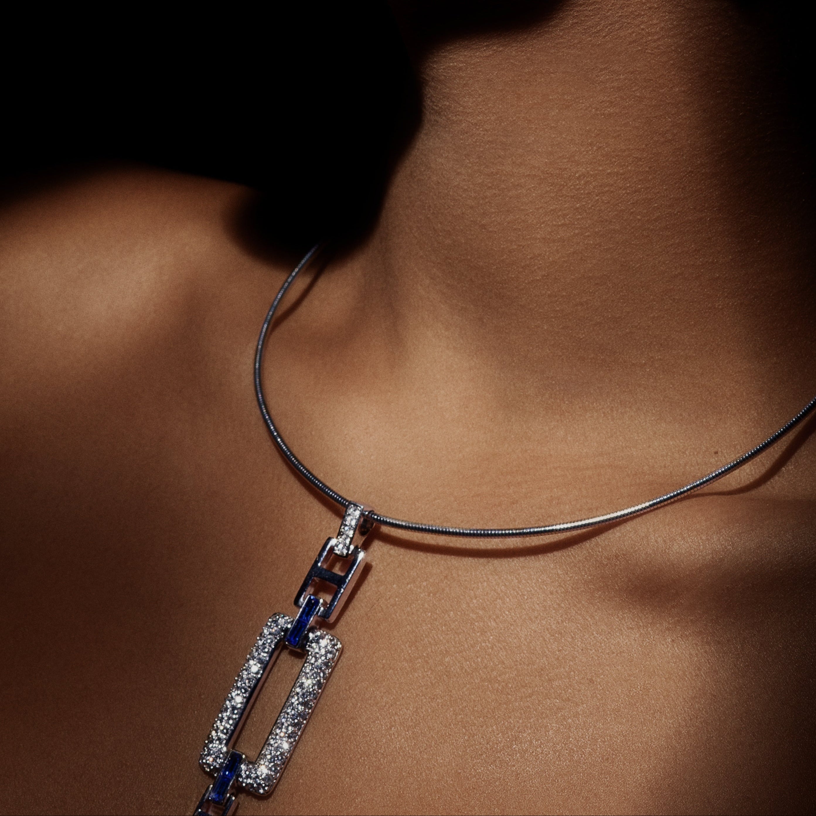 A close-up portrait of a woman wearing a silver-toned necklace and matching rectangular stud earring, both set with blue stones and clear pavé accents, against a dark background.