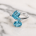A silver ring featuring a large emerald-cut light blue gemstone, a smaller round blue stone, and an oval light blue stone, displayed on a white marble surface.